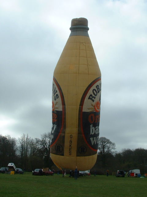 Side view of yellow balloon in the shape of a bottle with a green cap and the Robinsons Orange Barley Water label on the back and front.