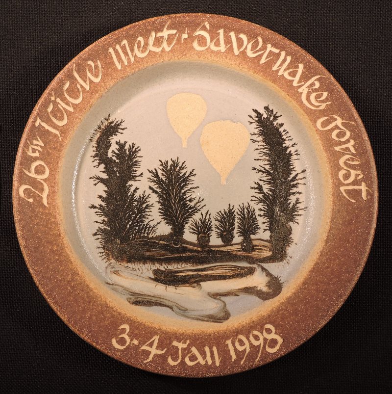 White ceramic plate with beige rim with event details in cream lettering. Centre of plate has 5 trees of varying size created in brown paint, with brown landscape. Silhouettes of 2 cream hot air balloons are flying above the trees.