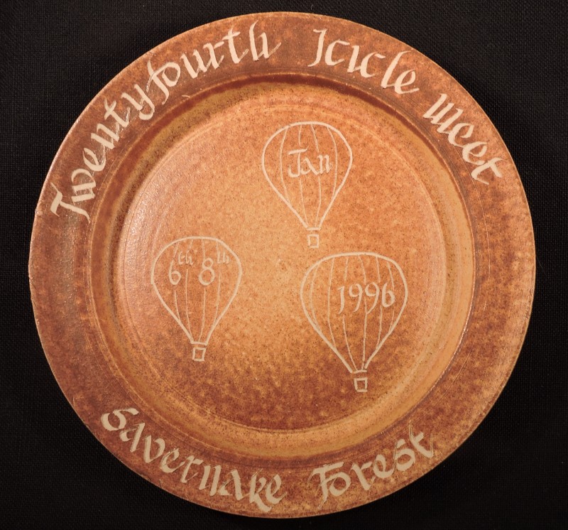 Light brown mottle effect ceramic plate, event details in cream on the rim. In the centre are three cream balloon line drawings, one has the event month, next has the event days, and last the year.