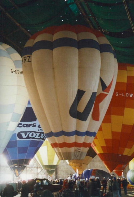 White balloon with a red, white and blue horizontal stripe at the top and base. Air UK logo in black and red in the centre white section. Photographed inflated with several other balloons inside a Cardington hanger.