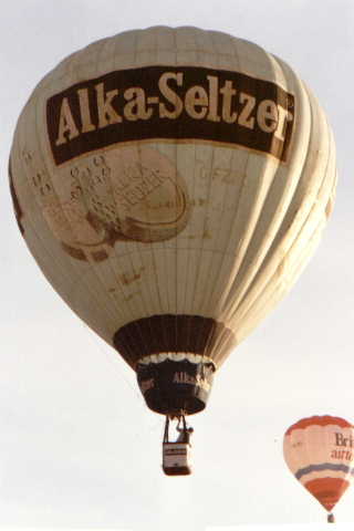 Light blue balloon in flight, with Alka Seltzer in white lettering in a black rectangle below which are two large Alka Seltzer tablets partly dissolved and fizzing.