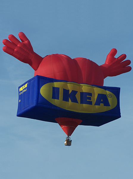 Photographed in a clear blue sky, the balloon is a blue rectangle box, with the IKEA logo, and a red heart, almost the length of the box, protruding from the top, with a red hand on each side of the heart. The base of the heart is below the box.