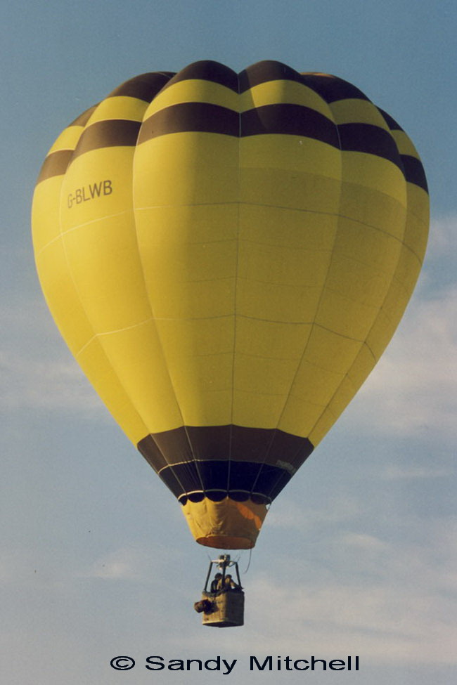 Yellow balloon in flight, has 2 brown stripes at the top and a brown and a black stripe at the mouth. 