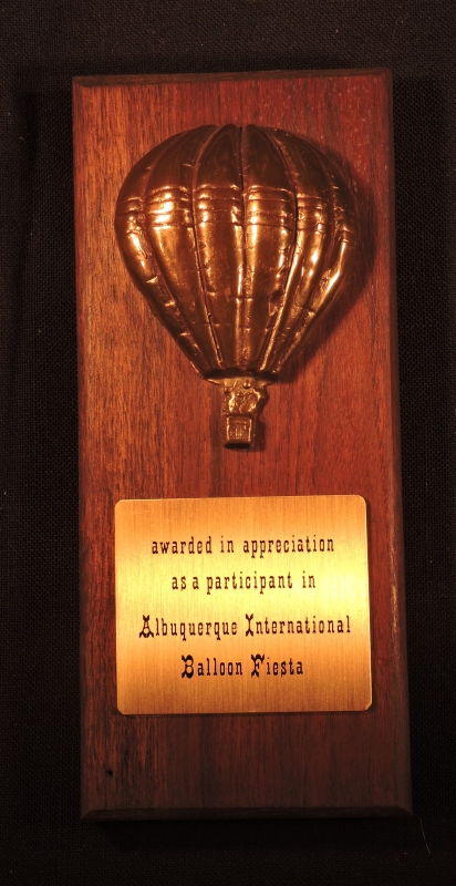 Upright rectangle dark wooden plaque. At top is a 3D metal image of balloon the same shade as the dark wood. Below is a gold-coloured metal plate with the words ‘awarded in appreciation as a participant in Albuquerque International Balloon Fiesta’.