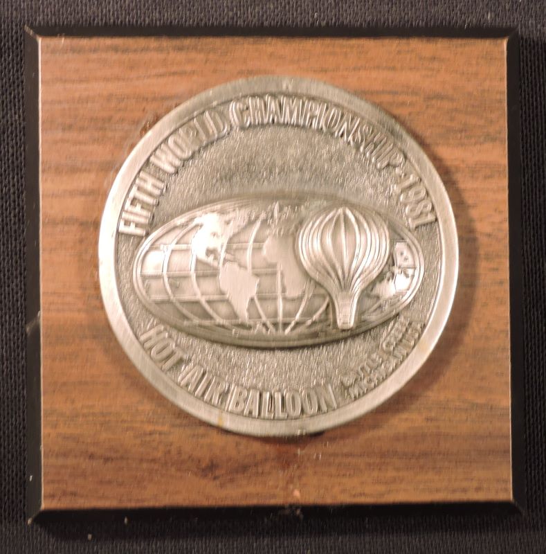 Round silver coloured metal plaque on square wooden base. In the centre of the plaque is an image of the world with a hot air balloon covering the area to the right of Africa. Event details around the edge of the plaque.