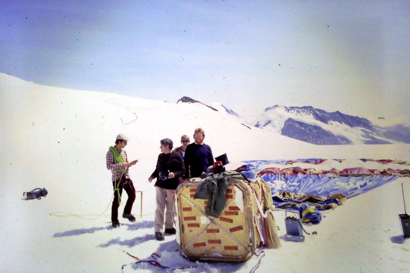 Colour picture of deflated balloon lying flat on snow on a mountain top with people standing beside the basket and mountain peak in the background.