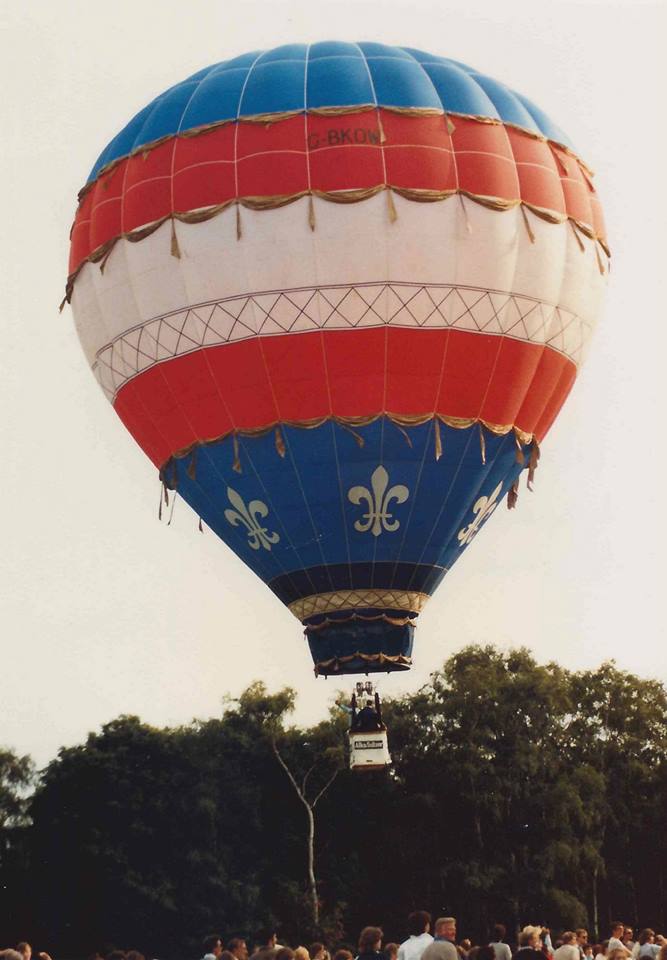Balloon airborne with horizontal stripes in blue, red, white, red, blue with fleur de lys motifs around lower blue panel and the name ELLE in black on the white centre panel.