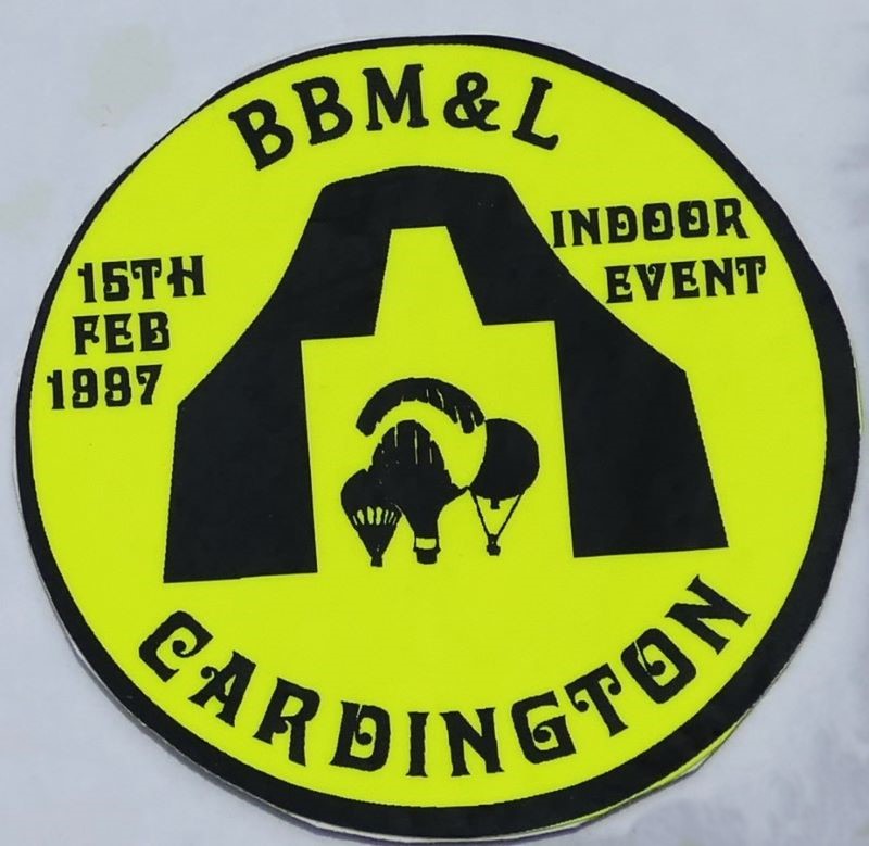 Yellow background to sticker with a black silhouette of 3 balloons in the entrance to the hangar in the centre. Event date and 