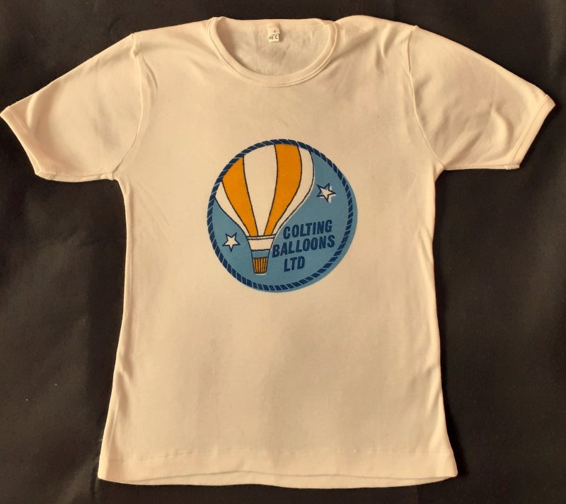 Off-white T-shirt with a light blue circle, edged in dark blue dashes, in the centre of the chest. Circle features a drawing of a yellow and white vertical striped balloon, the company name in dark blue and 2 white stars, edged in dark blue.