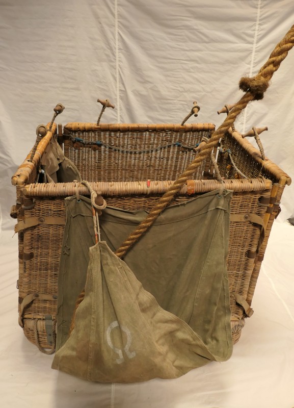 Square wicker basket with 2 wooden toggles on each side for attaching to the load ring. There is a heavy duty khaki fabric sack on one side, with the Omega symbol, which houses the trailrope. 
