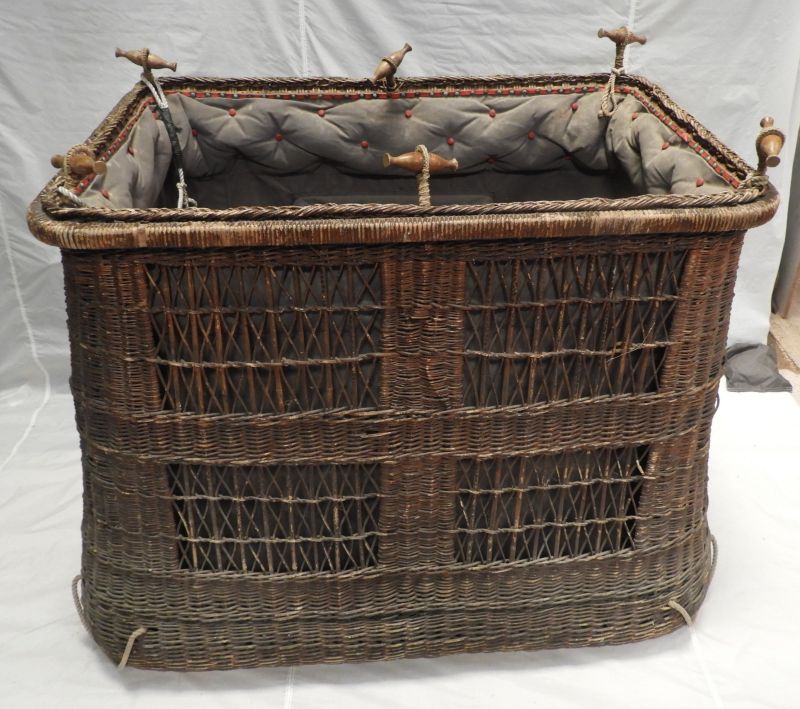 Wicker basket with 6 toggles to connect to load ring. Long sides have 4 rectangles of open wicker work divided into 9 rectangles across by 3 down with a wicker diagonal cross in each. The basket is lined with grey fabric, padded at the top edge.  