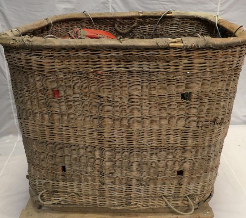 Wicker basket with 2 rope handles at base of each long side for carrying. The 4 gaps in the wicker are for the gas tank straps to pass through. Rim of the basket has a protective fabric covering. 8 metal flying wires are protruding from the rim.
