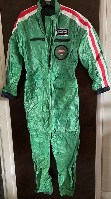 Green flying suit with white/red/white stripe along sleeves. Heineken name patch above Heineken balloon patch. Balloon is green with red horizontal stripe, white equator with 'Gerald A Heineken' in green, followed by red then green then white stripe.