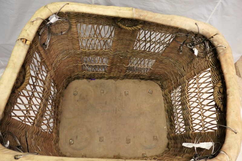 Looking into basket from above. There is a wooden floor. The end panels have 2 rectangles of open wicker work comprising wicker diagonal crosses. The 8 flying wires and 4 grab handles are visible.