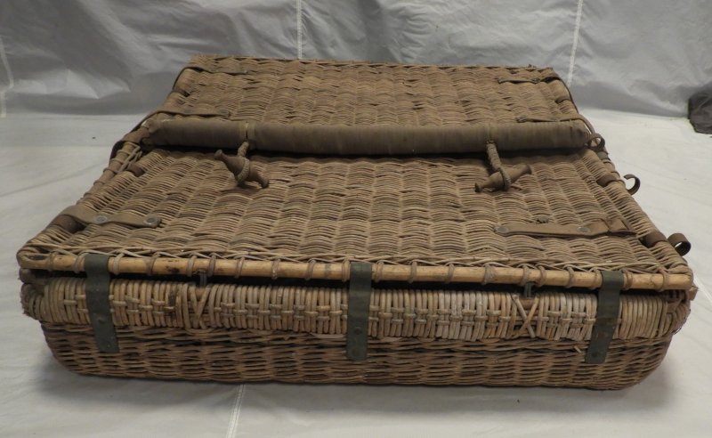 Wicker basket collapsed flat onto the wicker base. Each side has a cane frame with wicker woven on to it and has 3 metal retaining strips. The metal corner pole rings and leather tank straps are also visible.      