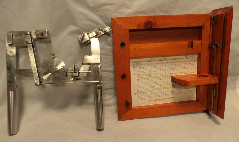  Silver coloured metal instrument with 2 handles with clip at top to hold the balloon fabric. The handles are joined by a bar, with round dial with the pressure readings, indicated by a needle. It is stored in a wooden box complete with instructions.      