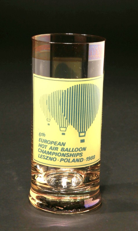 Tall glass with a feint gold band at the rim. 3 balloons silhouetted in blue vertical lines are one behind the other in increasing size on a large yellow square. The square has a blue line just inside the edge and includes the event details in blue.