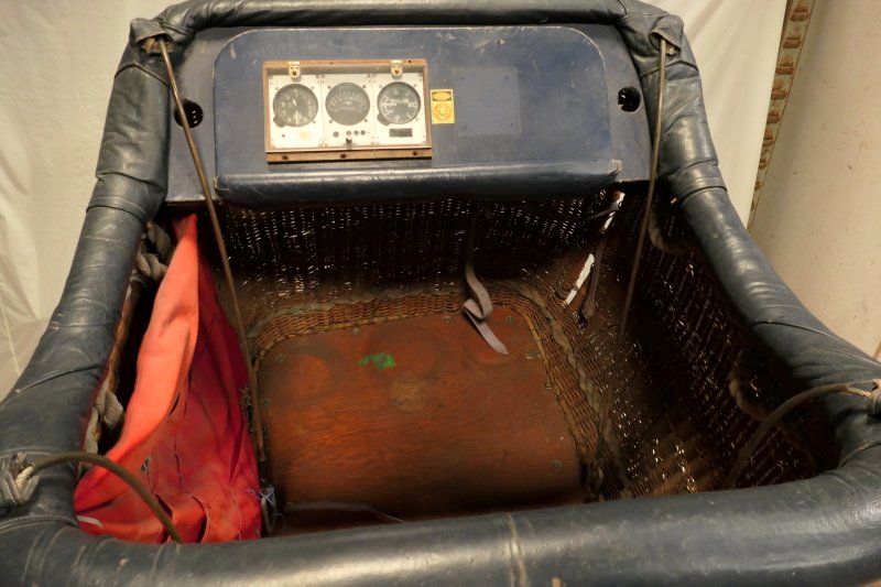 View looking down into the basket from the short edge showing the instrument panel across the opposite short edge, a red fabric bag with pockets on one long edge, the wooden floor and 4 flying wire are also visible.   
