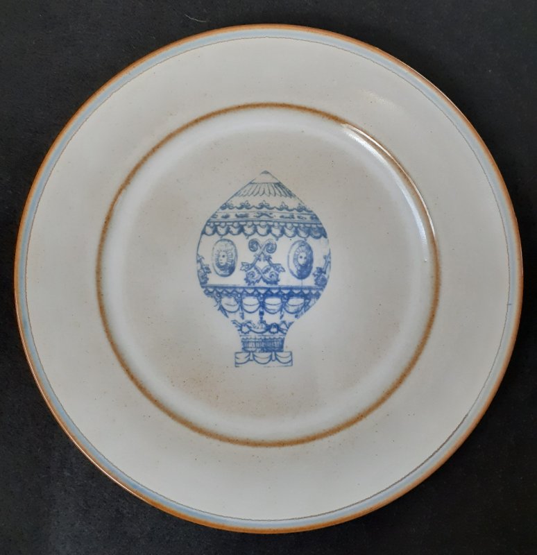 Brown earthenware plate, lightly painted in pale grey with earthenware slightly showing through. Both sides of wide rim are not painted. Outer rim has a pale blue and a brown line. In centre is pale blue drawing of the Montgolfier balloon.